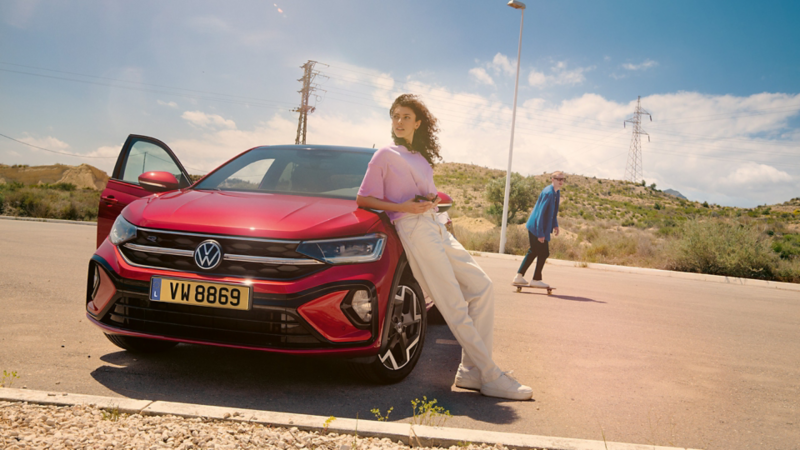 Une femme est appuyée sur le côté du capot d'une Volkswagen rouge. À l'arrière-plan, un homme fait du skateboard.