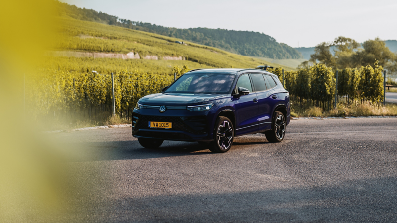 Un SUV Volkswagen Tayron bleu foncé est garé au milieu d'une route de campagne, avec des vignobles en arrière-plan. 