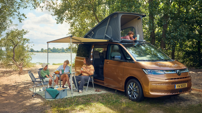 Van Volkswagen California Beach Tour stationné au bord d’un lac, toit relevable ouvert, avec des personnes installées sous un auvent à côté d’un espace pique-nique.