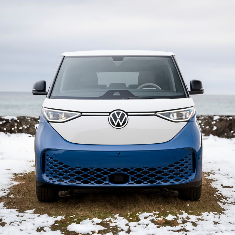 A blue and white 2025 VW ID. Buzz parked on a road with hills and houses in the background.