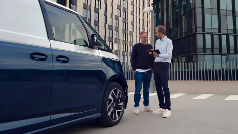 VW Nutzfahrzeug auf einem Parkplatz vor modernen Bürogebäuden. Zwei Personen stehen neben dem Fahrzeug und betrachten gemeinsam ein Tablet.