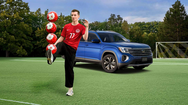 Pier-Luc Funk porte un maillot de Canada Soccer tout en maintenant en équilibre sur son pied une pile verticale de quatre ballons de Canada Soccer. Il se tient debout sur un terrain de soccer en pelouse synthétique verte à côté d'un Volkswagen Atlas, avec un but de soccer et des arbres visibles à l'arrière-plan sous un ciel nuageux.
