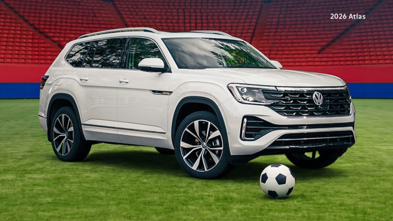 A Volkswagen Atlas is on a green soccer field, with a soccer ball placed near the front of the car. In the background are rows of empty red stadium seats and red-and-blue banners.