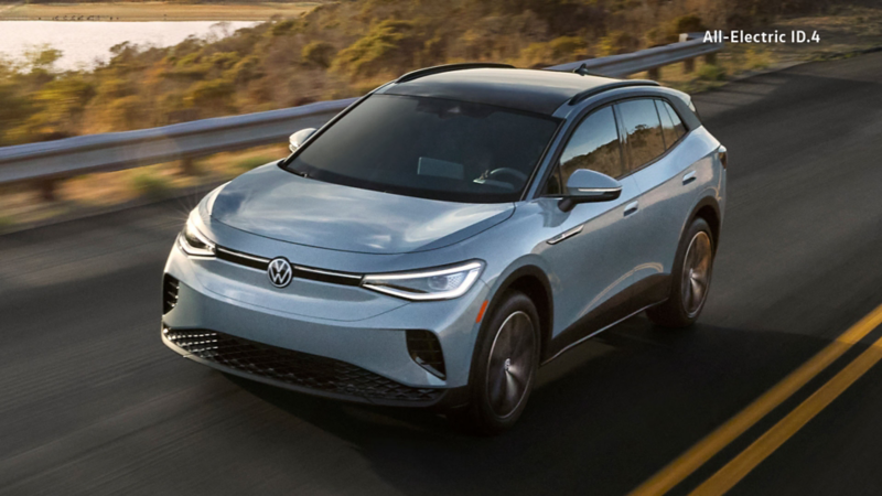 An Ice Grey Metallic ID.4 Pro S AWD, driving on a paved road with a lake, trees, and guardrail in the background during daylight.
