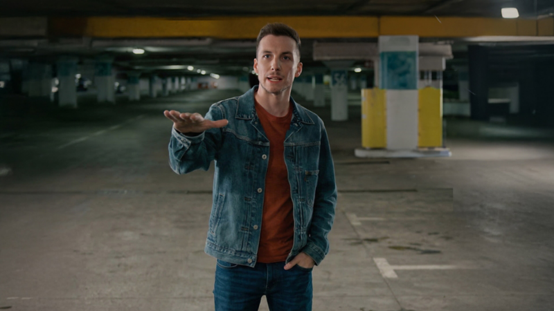 Pier-Luc Funk se tient dans un stationnement souterrain vide, face à la caméra, avec une main tendue vers l’avant. Il porte une veste en denim par-dessus un chandail couleur rouille et un jean. L’espace est faiblement éclairé, avec des piliers en béton, des accents jaunes peints et des rangées de places de stationnement qui s’étendent à l’arrière-plan.