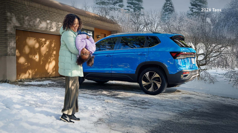 A woman holding her daughter in a snowy driveway and both of them are smiling. There’s a blue Taos parked in the background.