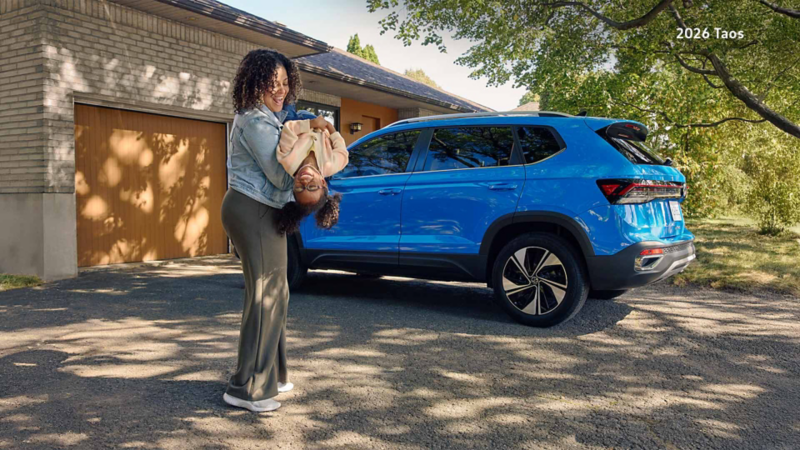 A woman holds a young girl upside down, both smiling, next to a Cornflower Blue Volkswagen Taos parked in a driveway.