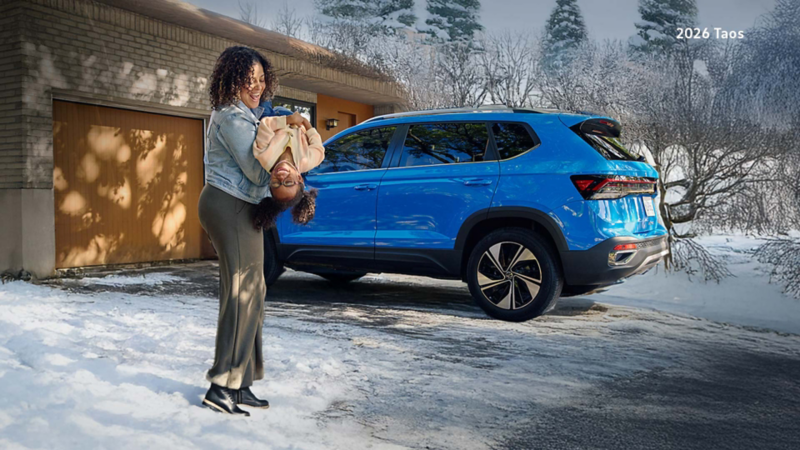 A woman holding her daughter in a snowy driveway and both of them are smiling. There’s a blue Taos parked in the background.