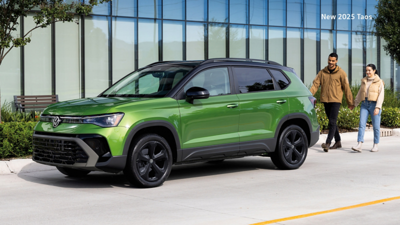 A Serrano Green Metallic Volkswagen Taos Comfortline Black Edition parked on a modern city street in front of a building with large glass windows, as a smiling couple walks by on the sidewalk.