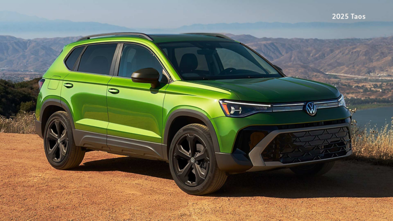 A Serrano Green Metallic Volkswagen Taos Comfortline Black Edition parked on a scenic dirt overlook with rolling hills and a lake in the background.