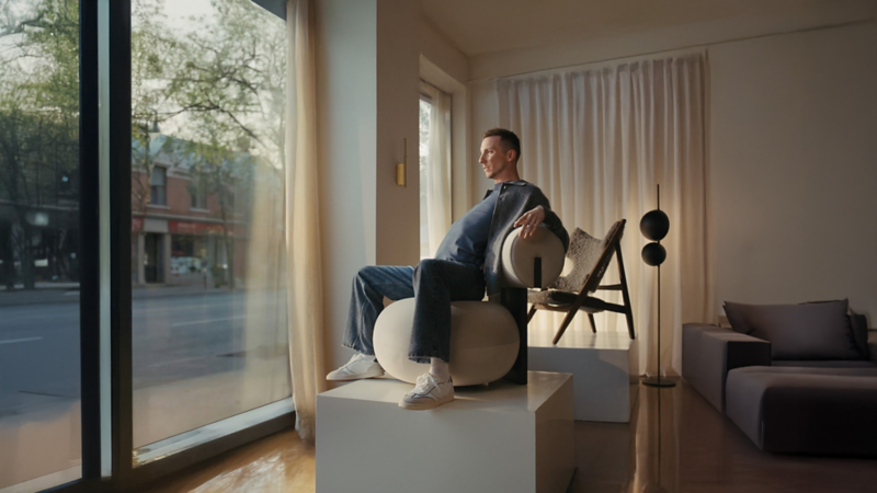 Pier-Luc Funk est assis sur un fauteuil moderne aux formes arrondies, placé sur un socle près d’une grande fenêtre laissant entrer une douce lumière naturelle. Il porte un jean, des espadrilles blanches et un haut foncé, regardant vers la rue. L’intérieur est minimaliste, avec des rideaux clairs, un plancher en bois et une lampe contemporaine en arrière-plan.