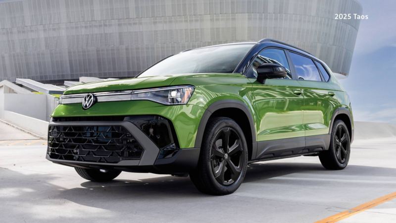 A Serrano Green Metallic Taos Comfortline Black Edition, parked on a concrete surface in front of a modern architectural building under a clear sky.