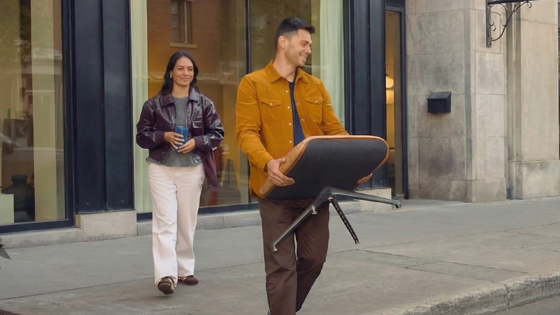 Un homme sort d’un immeuble moderne en ville en portant une chaise, tandis qu’une femme le suit sur le trottoir avec une tasse à la main. Les bâtiments en pierre et en brique bordent la rue calme, avec quelques arbres et vitrines en arrière-plan.