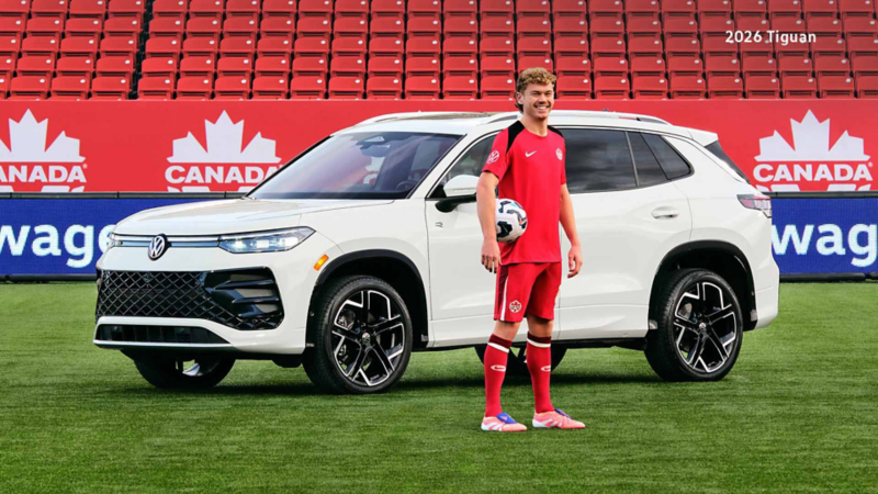 Jacob Shaffelburg is in a Team Canada Soccer uniform on a green field holding a soccer ball, posed in front of a Tiguan. Behind him are rows of empty red stadium seats and banners featuring the Canada Soccer logo and Volkswagen branding.