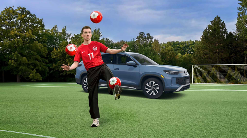 Pier-Luc Funk porte un maillot de Canada Soccer tout en jonglant avec trois ballons de Canada Soccer sur un terrain en gazon synthétique vert. Il est en pleine action, maintenant un ballon en équilibre sur son pied tandis que deux autres flottent dans les airs autour de lui. Derrière lui se trouve un Volkswagen Tiguan, avec un but de soccer et des arbres visibles à l'arrière-plan sous un ciel nuageux.