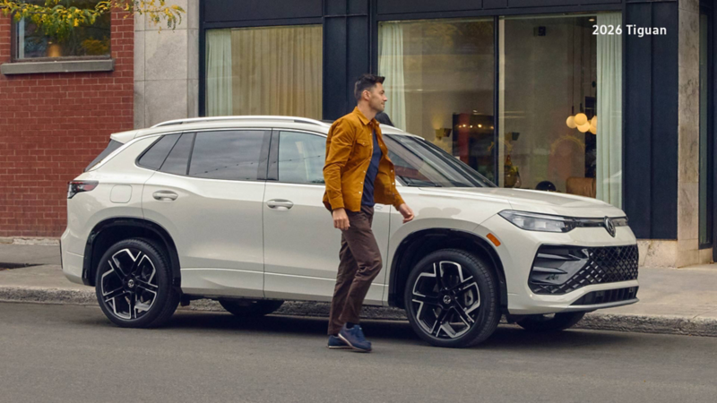 A man in a brown jacket walks alongside an Ascot Grey Volkswagen Tiguan parked on a city street in front of a building with brick and modern facades.