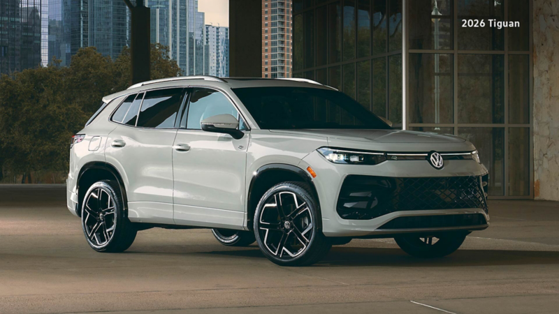 A Volkswagen Tiguan is parked on a light gray concrete floor within a modern, open-air structure. In the background, a cityscape with tall buildings and some trees is visible under a light sky.