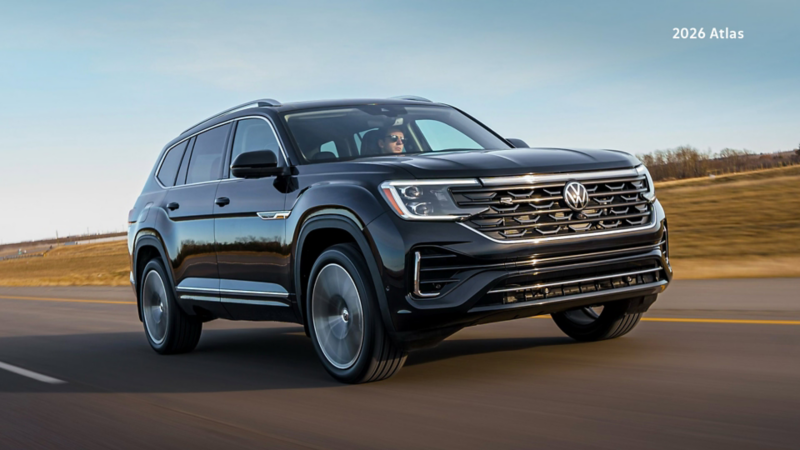 A Volkswagen Atlas is in motion on a multi-lane highway. The highway curves slightly to the right and is surrounded by a light brown grassy landscape and a clear blue sky. There is motion blur indicating movement.