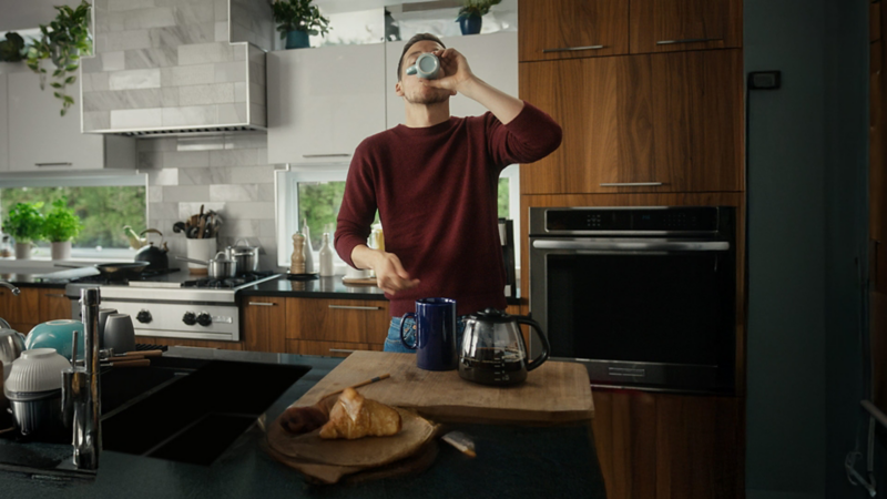 Pier-Luc Funk dans une cuisine, avec une cuisinière, des casseroles et un four encastré visibles en arrière-plan. Il porte un chandail à manches longues et semble préparer de la nourriture sur un comptoir.