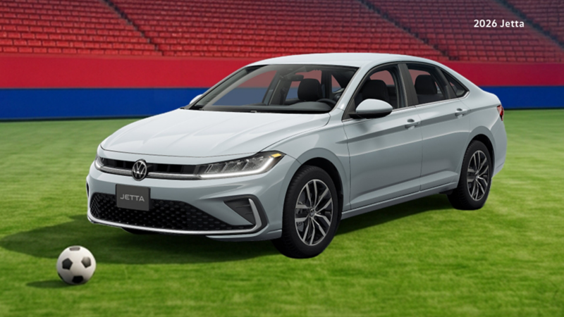 A Volkswagen Jetta is on a green soccer field, with a soccer ball placed near the front of the car. In the background are rows of empty red stadium seats and red-and-blue banners. 