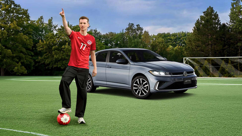  Pier-Luc Funk porte un maillot de Canada Soccer alors qu'il se tient debout sur un terrain vert, un pied posé sur un ballon de soccer, tout en pointant vers le lointain. À côté de lui se trouve une Volkswagen Jetta, avec un but de soccer et des arbres visibles à l'arrière-plan sous un ciel nuageux.
