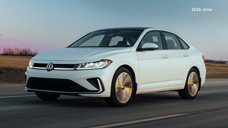 An Oryx White Pearl Volkswagen Jetta drives on a highway at dawn, with a blue and pink sky above a flat, brown landscape.