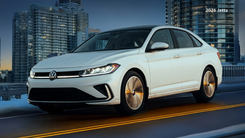 A white Jetta is driving on a road alongside under cloudy skies. City buildings are seen in the background.
