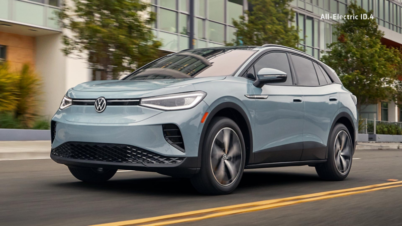 An Ice Grey Metallic Volkswagen ID.4 Pro S driving on a city street near modern buildings and greenery. 