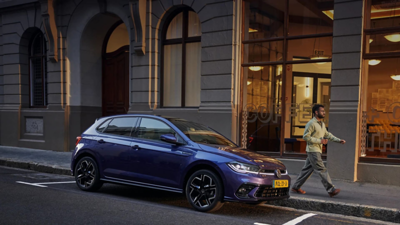 Een blauwe Volkswagen Polo staat geparkeerd langs een stoep naast een horecagelegenheid. Een man loopt weg van de auto