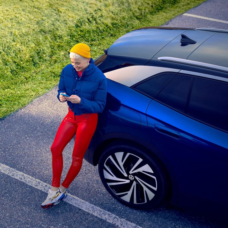 Vue oblique du hayon d’une VW ID.4 bleue, une personne habillée en vêtements de sport s’appuie sur la voiture et utilise son smartphone