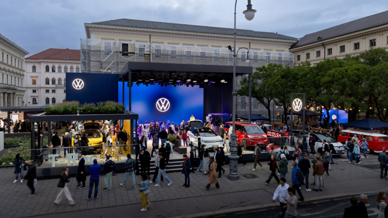 Volkswagen exhibition stand outdoors with a stage, several cars, and many visitors.