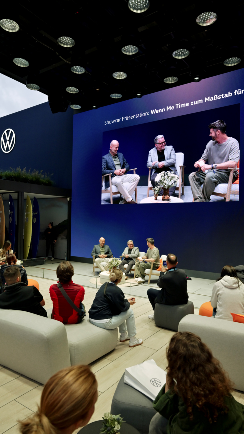Visitors seated on lounge furniture watching a talk on the large Volkswagen stage with screen.