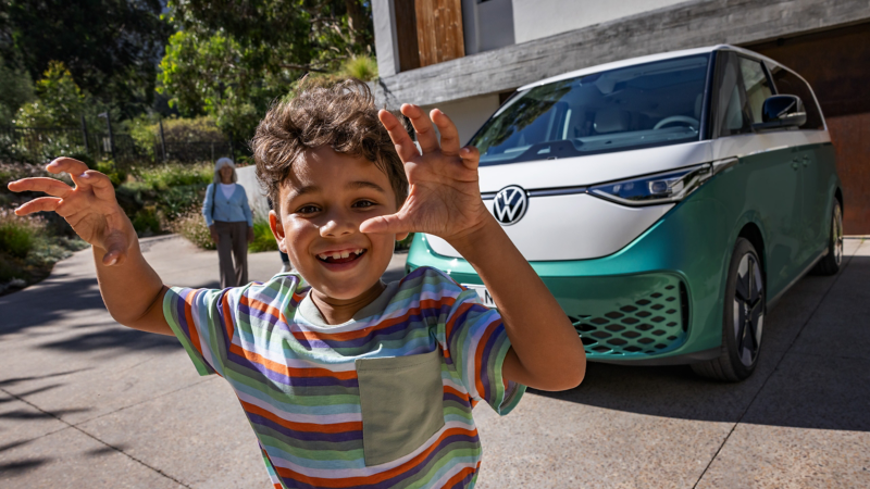 Ein VW ID. Buzz steht auf einer Auffahrt vor einem Haus. Im Vordergrund steht ein Kind vor der Kamera und hält in einer spielerischen Geste die Hände nach oben.