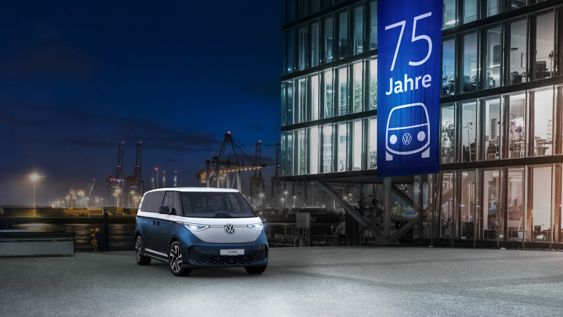 Ein blauer und weißer VW ID. Buzz steht nachts vor einem beleuchteten Gebäude mit einem großen Banner zum "75 Jahre" Jubiläum.