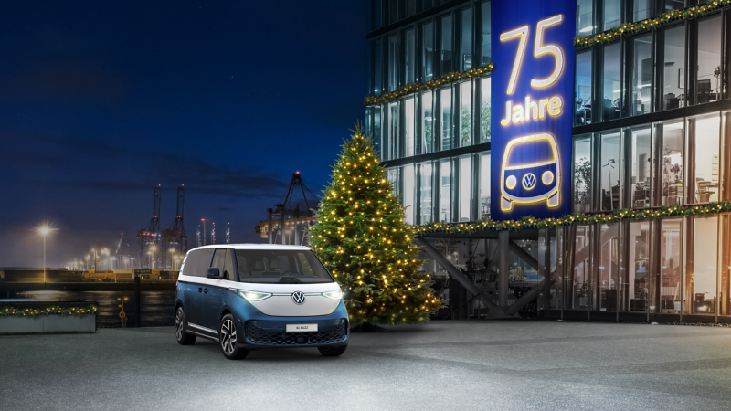 Ein blauer und weißer Volkswagen ID. Buzz steht nachts vor einem festlich beleuchteten Gebäude mit einem großen Banner zum "75 Jahre" Jubiläum und einem Weihnachtsbaum davor.