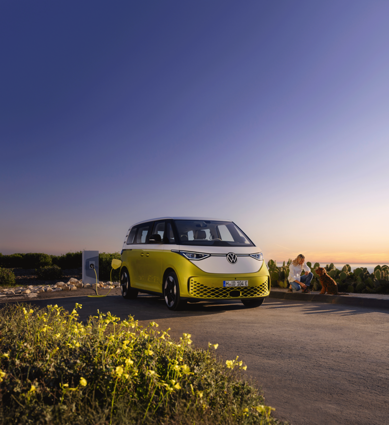 At dusk on the beach, a woman is petting a dog, a VW ID. Buzz is parked next to it.