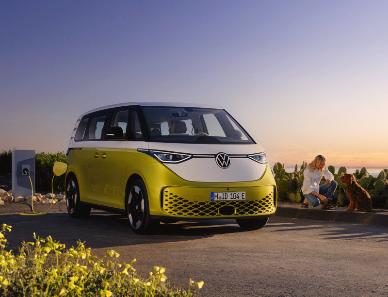 W blasku zachodzącego słońca, kobieta głaszcze psa przy plaży, obok stoi VW ID. Buzz.