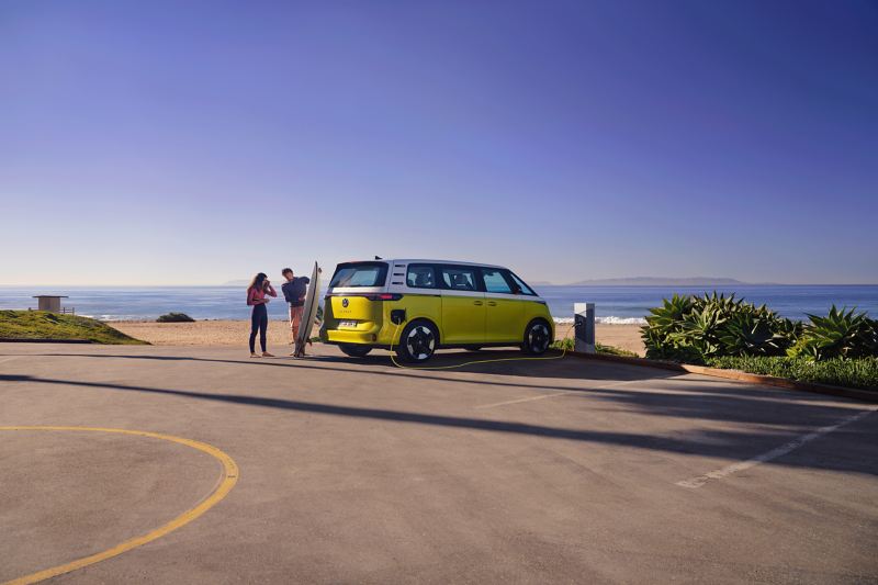 The VW ID. Buzz parks on the beach, in front of it a man shows his daughter a surfboard.