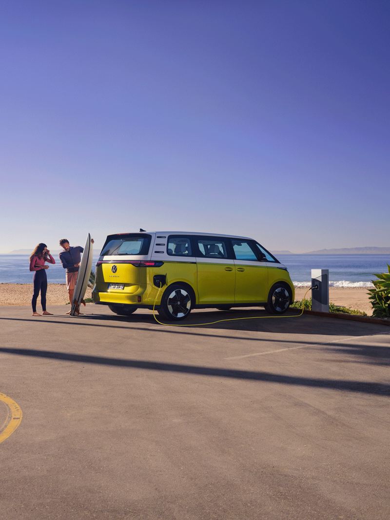 L’ID. Buzz VW est garée à la plage, devant le véhicule, un homme montre une planche de surf à sa fille.