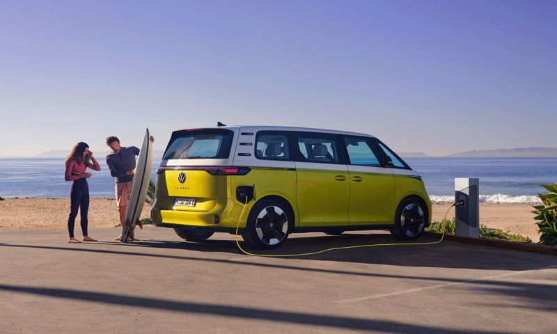 VW ID. Buzz parkerad på stranden och framför den står en man och visar sin dotter en surfbräda.