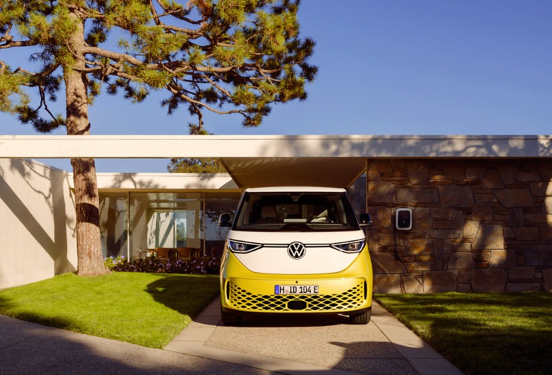 Un Volkswagen ID.Buzz color giallo-bianco fermo sul lungomare. Sullo sfondo, una famiglia con due bambini ammira il tramonto sul mare. 