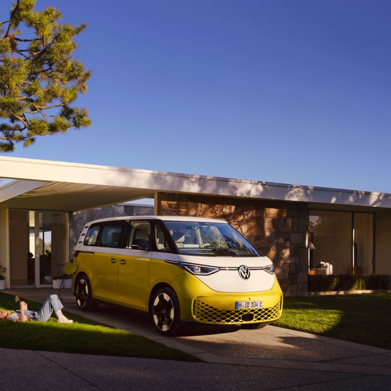 Ein weiß-gelber VW ID. Buzz steht auf einem Platz, umgeben von einer Familie. Front und Seite des Fahrzeugs sichtbar.