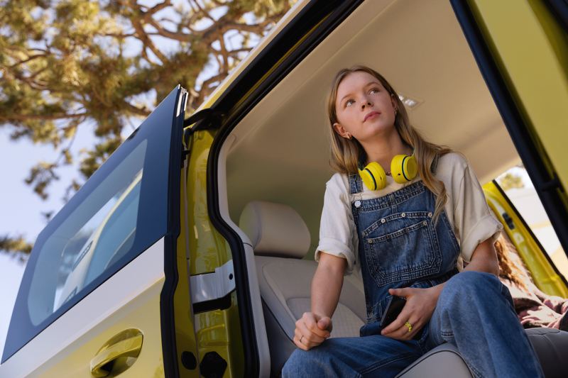 Dziewczynka z żółtymi słuchawkami siedzi w bocznych rozsuwanych drzwiach VW ID. Buzz.