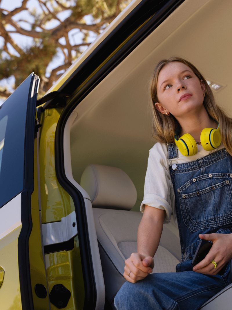 Jeune fille avec avec un casque audio jaune est assise sur le siège arrière d'un VW ID. Buzz jaune et blanc par l'ouverture de la porte.