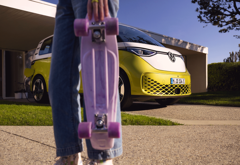 The ID. Buzz stands in a driveway, with a girl holding a purple skateboard in the foreground. 