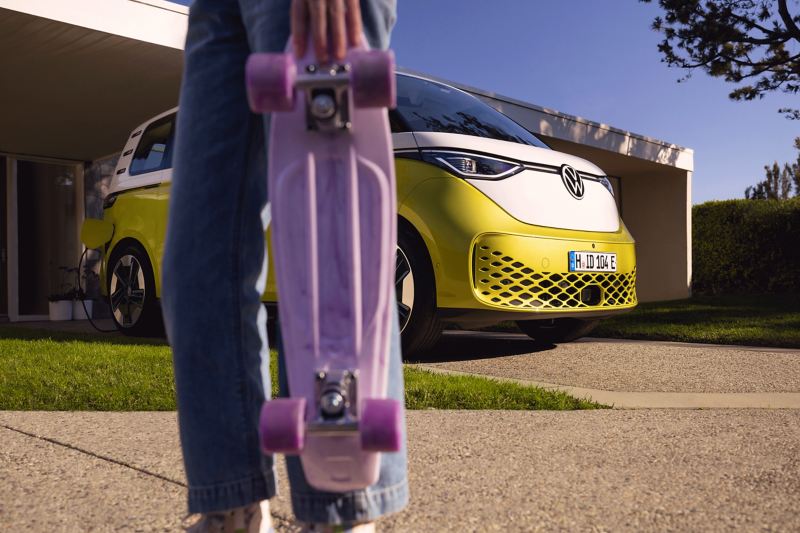 The ID. Buzz stands in a driveway, with a girl holding a purple skateboard in the foreground. 