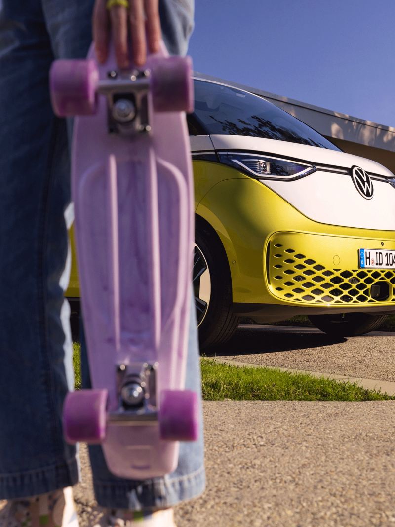 L'ID. Buzz jaune et blanc stationné devant une maison avec un skateboard violet au premier plan.