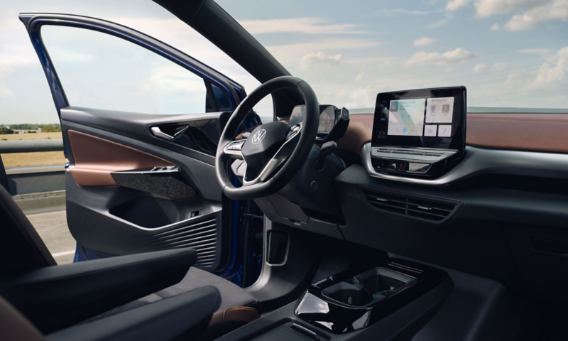 Interior view of the cockpit in the VW ID.4 with multifunction steering wheel, centre console and touch display. View from windscreen to blue sky, with door open.