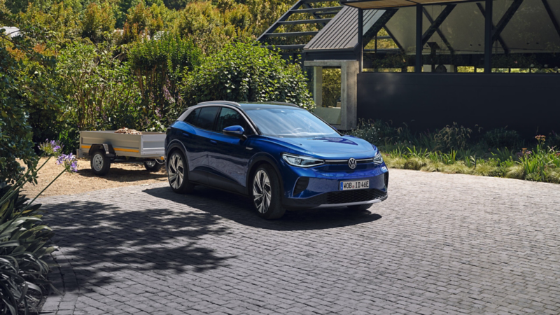 VW ID.4 in blau von vorne und Seite auf Hof stehend. Ein Anhänger ist angeschlossen.