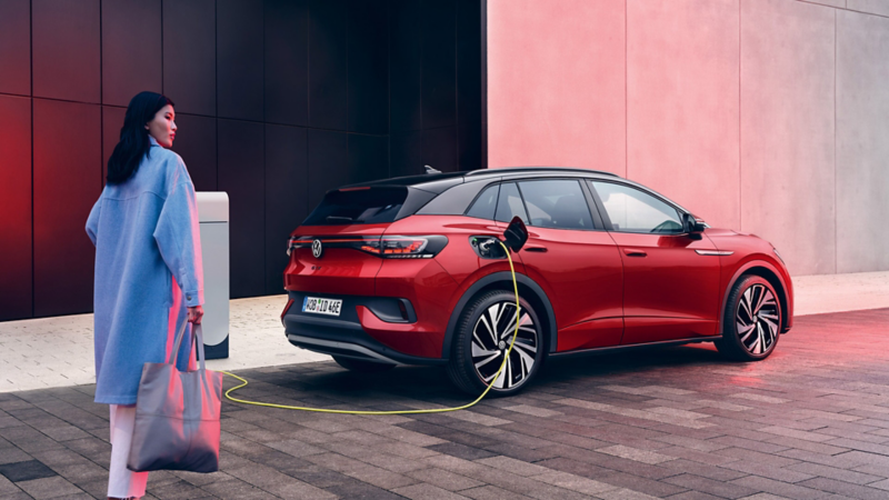 Red Volkswagen electric ID.4 GTX charges at a wall-mounted station as a person arrives with shopping outside a modern building.